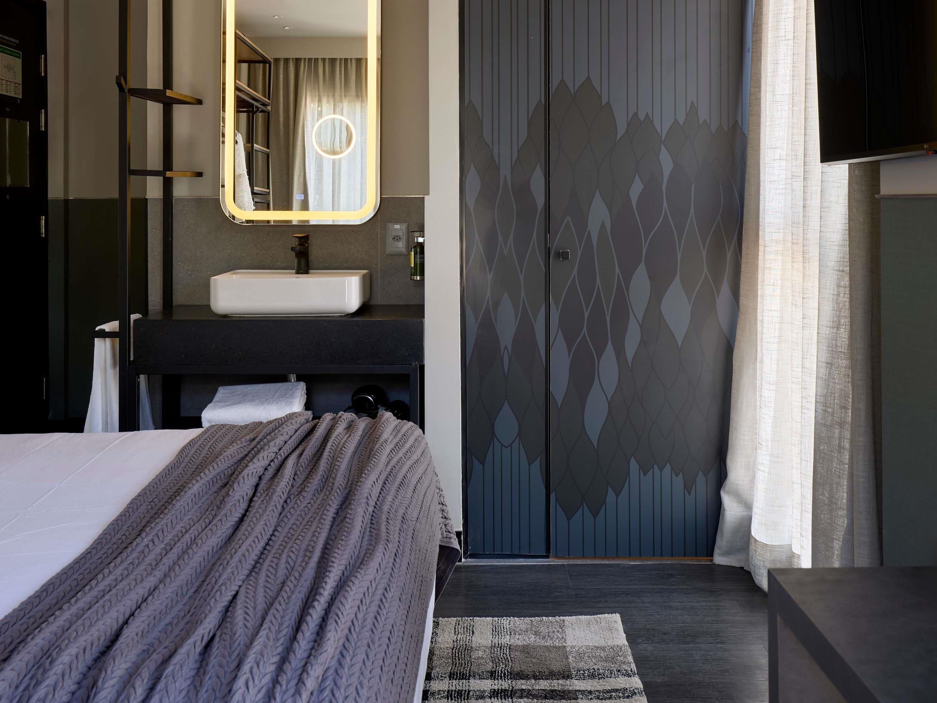 Una habitación de hotel moderna con una cama, un baño con lavamanos y espejo, y un armario.