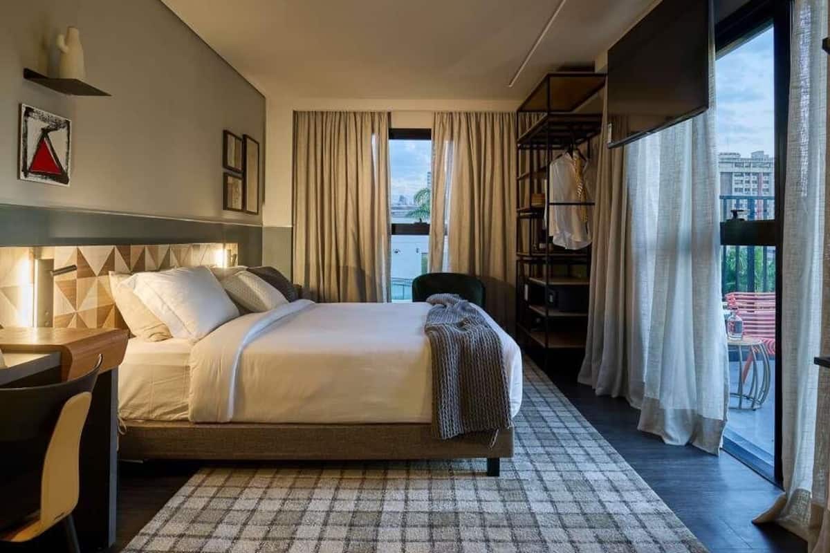 Una habitación de hotel moderna con una cama, un escritorio, una silla, una estantería y un ventanal con cortinas.