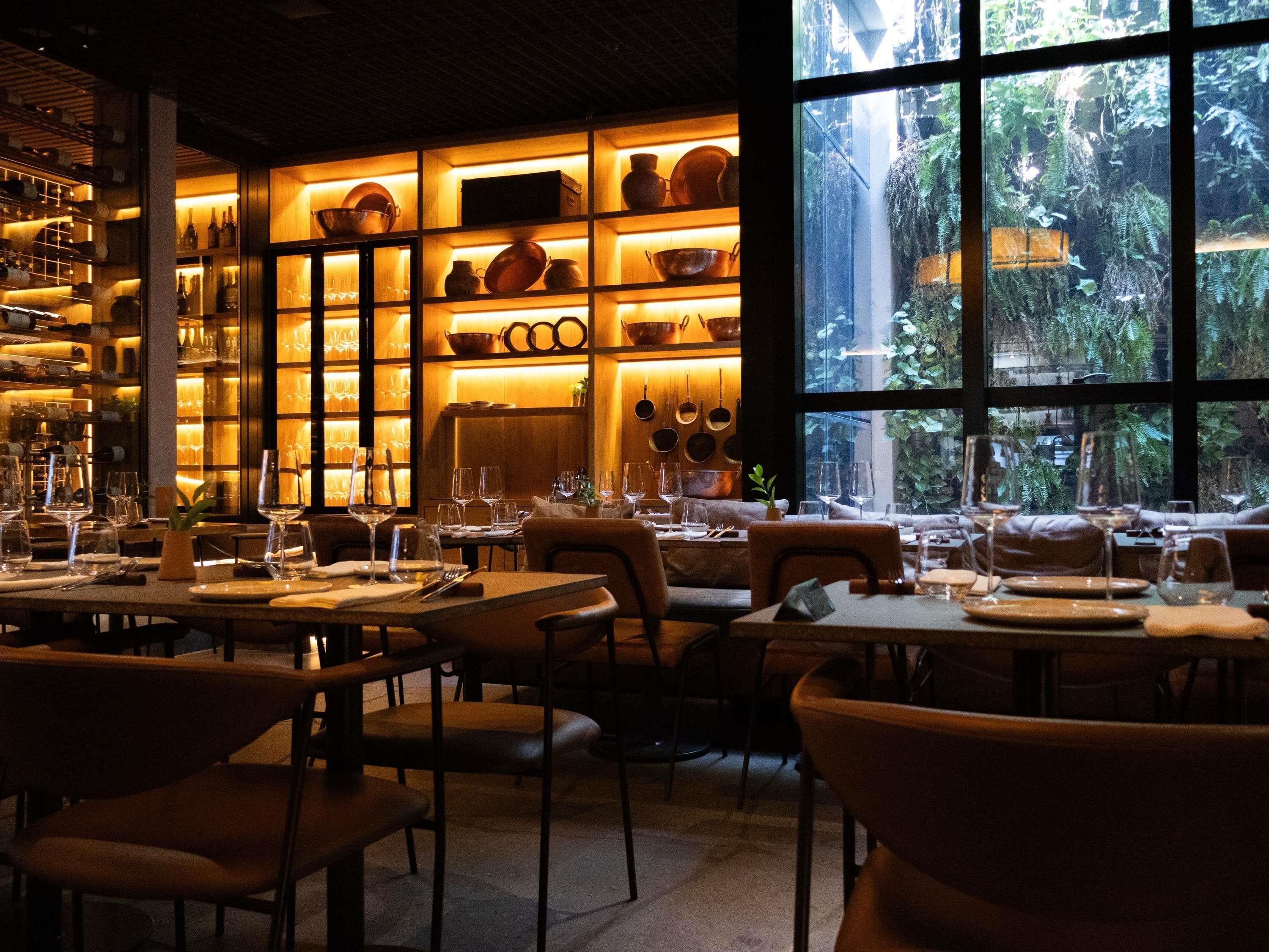Um restaurante com mesas preparadas para refeições, uma adega e vista para o verde através de grandes janelas.