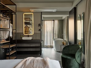 Habitación de hotel con cama, tocador con espejo, un sillón verde y vistas al exterior a través de amplias ventanas.