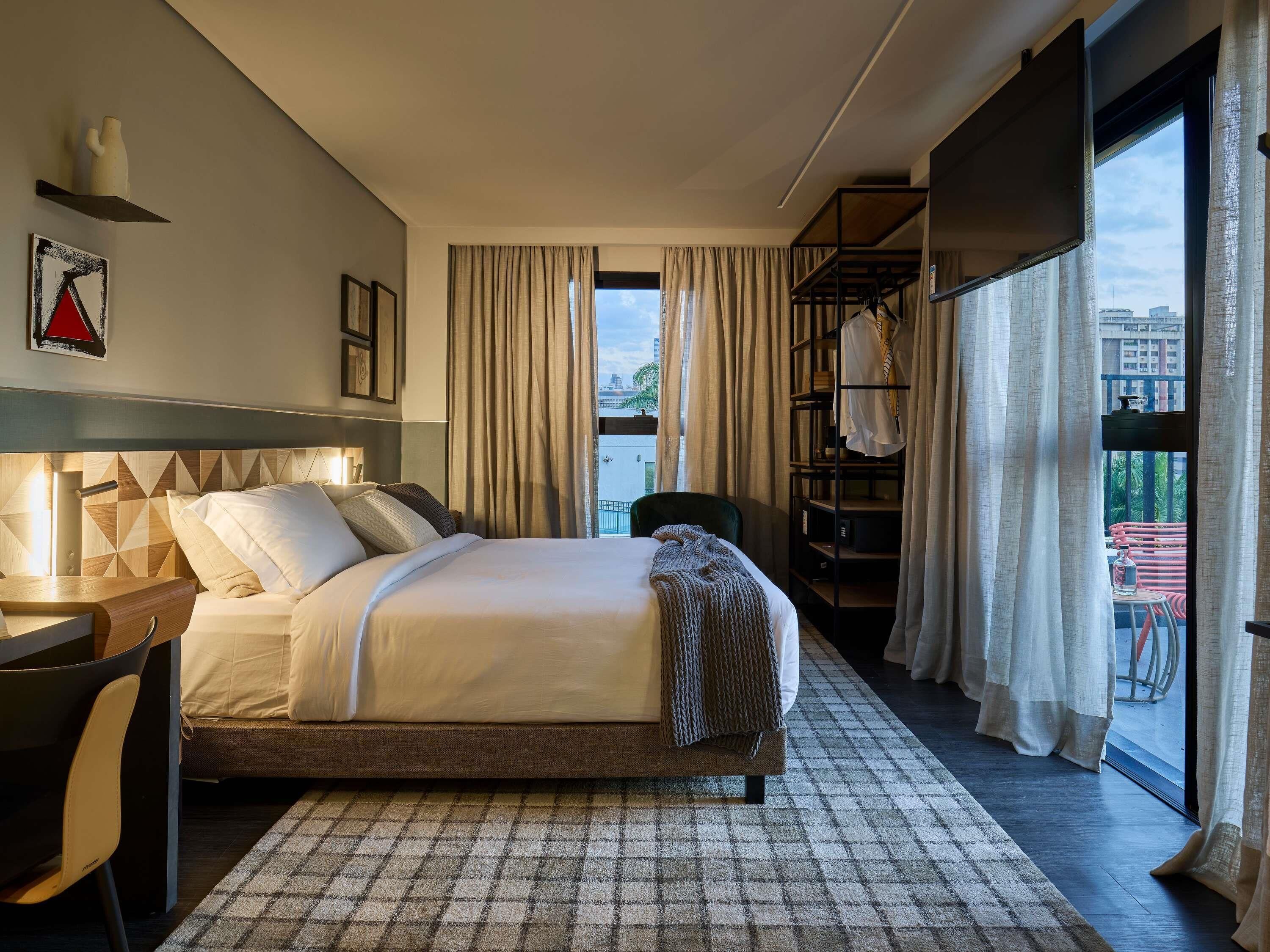 A modern hotel room with a bed, a desk, a chair, a bookshelf, and a view of the city.