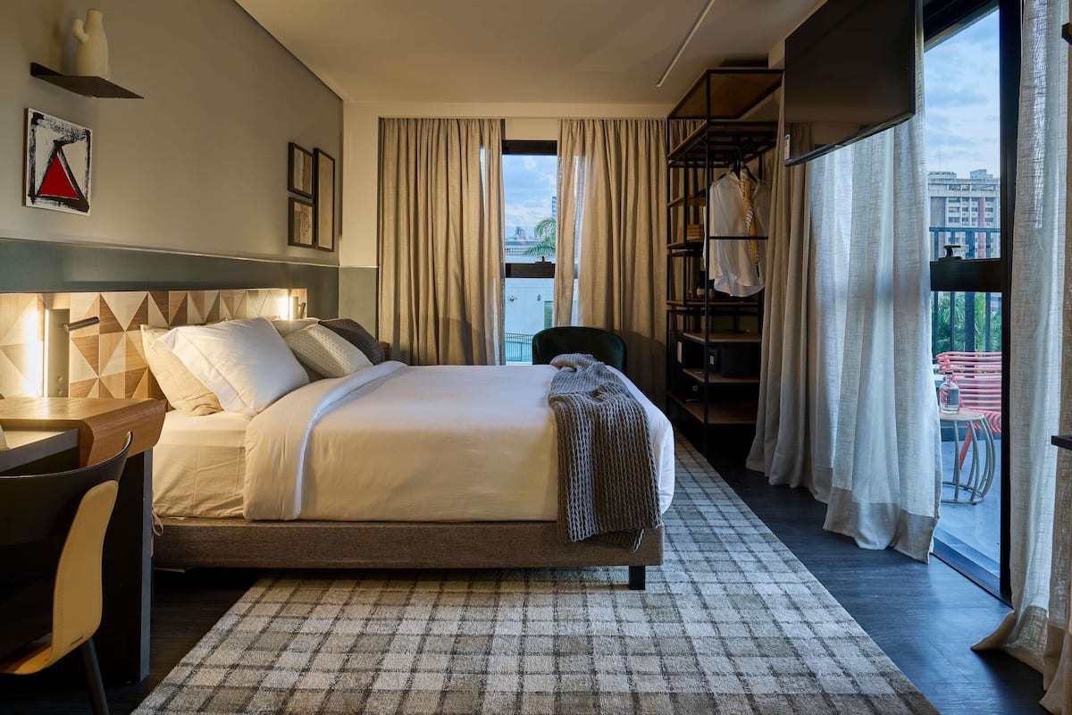 A modern hotel room with a bed, a desk, a chair, a bookshelf, and a view of the city.