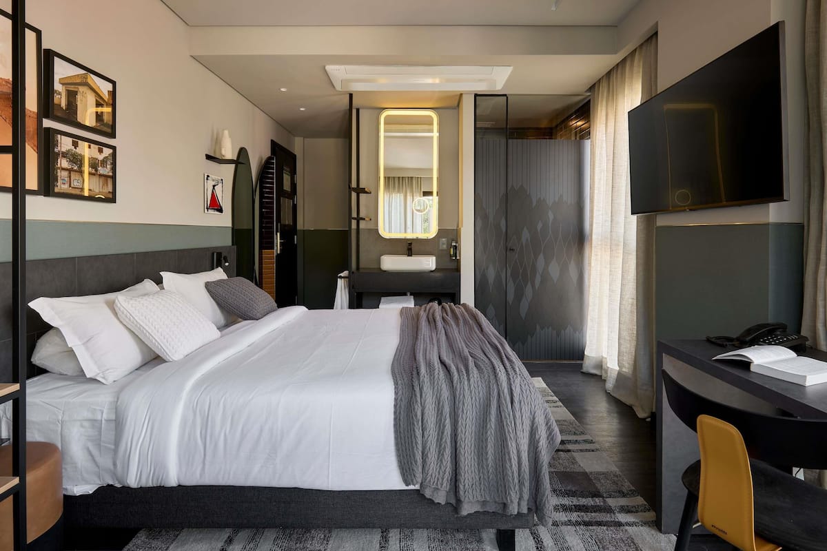 A modern hotel room with a large bed, a desk with a chair, a TV, and a bathroom visible through an open door.