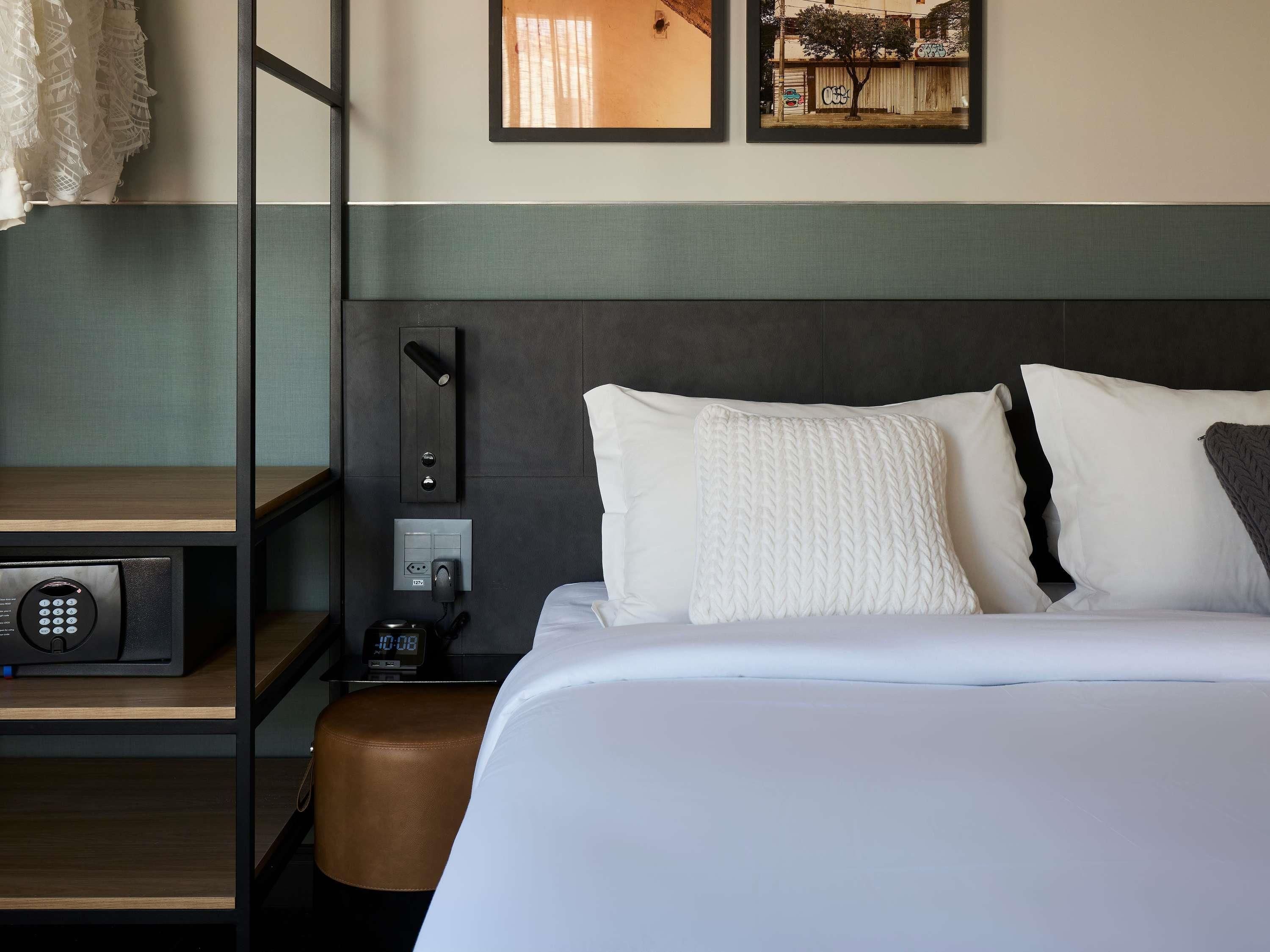 Habitación de hotel con una cama, una mesita de noche con un reloj digital, un enchufe y una lámpara de pared.