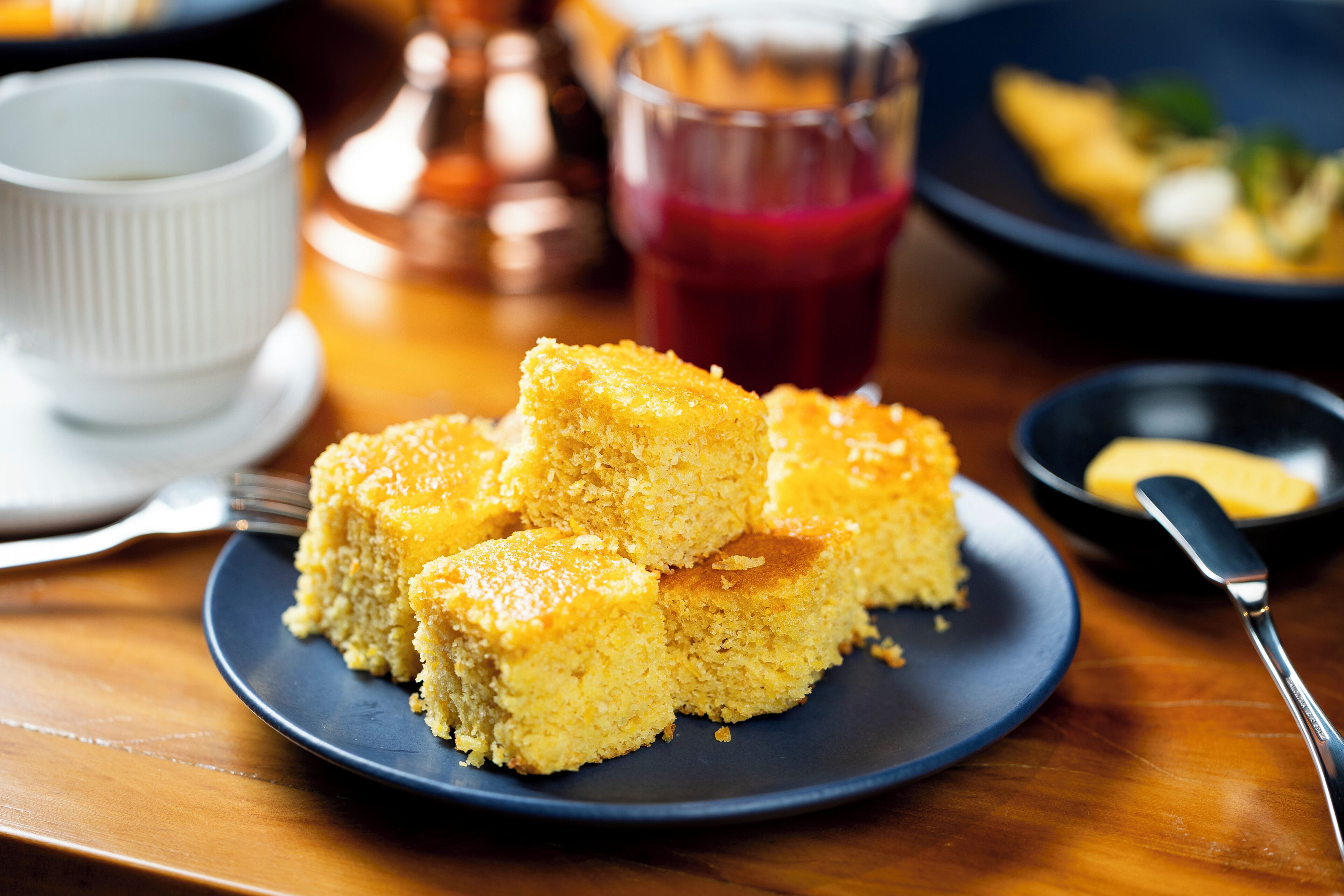 A plate of cornbread on a dark blue plate with a fork and a small bowl of butter on the side.