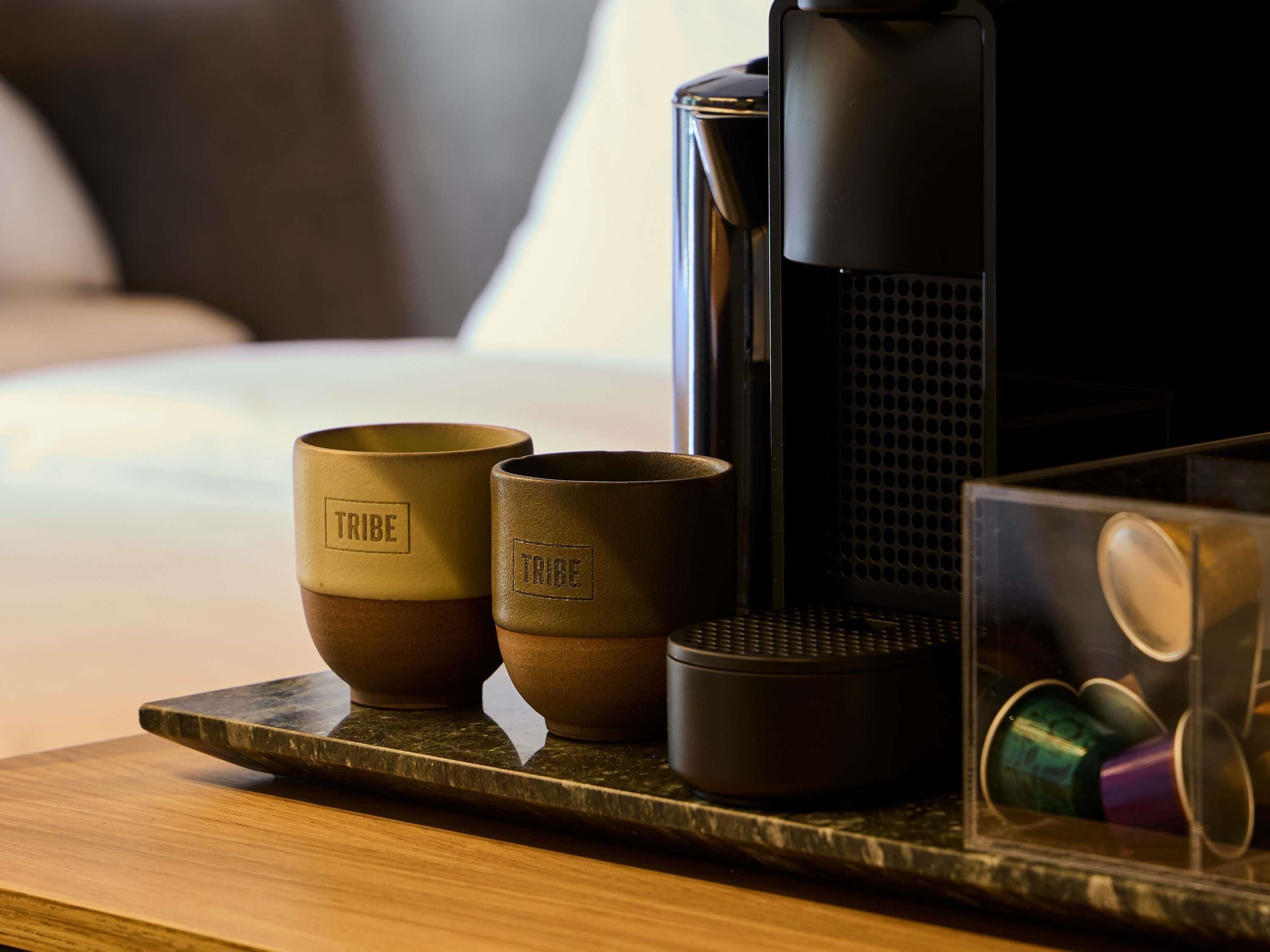 Una cafetera, dos tazas con el nombre 'TRIBE' grabado y un recipiente con tapas de colores sobre una superficie de madera.