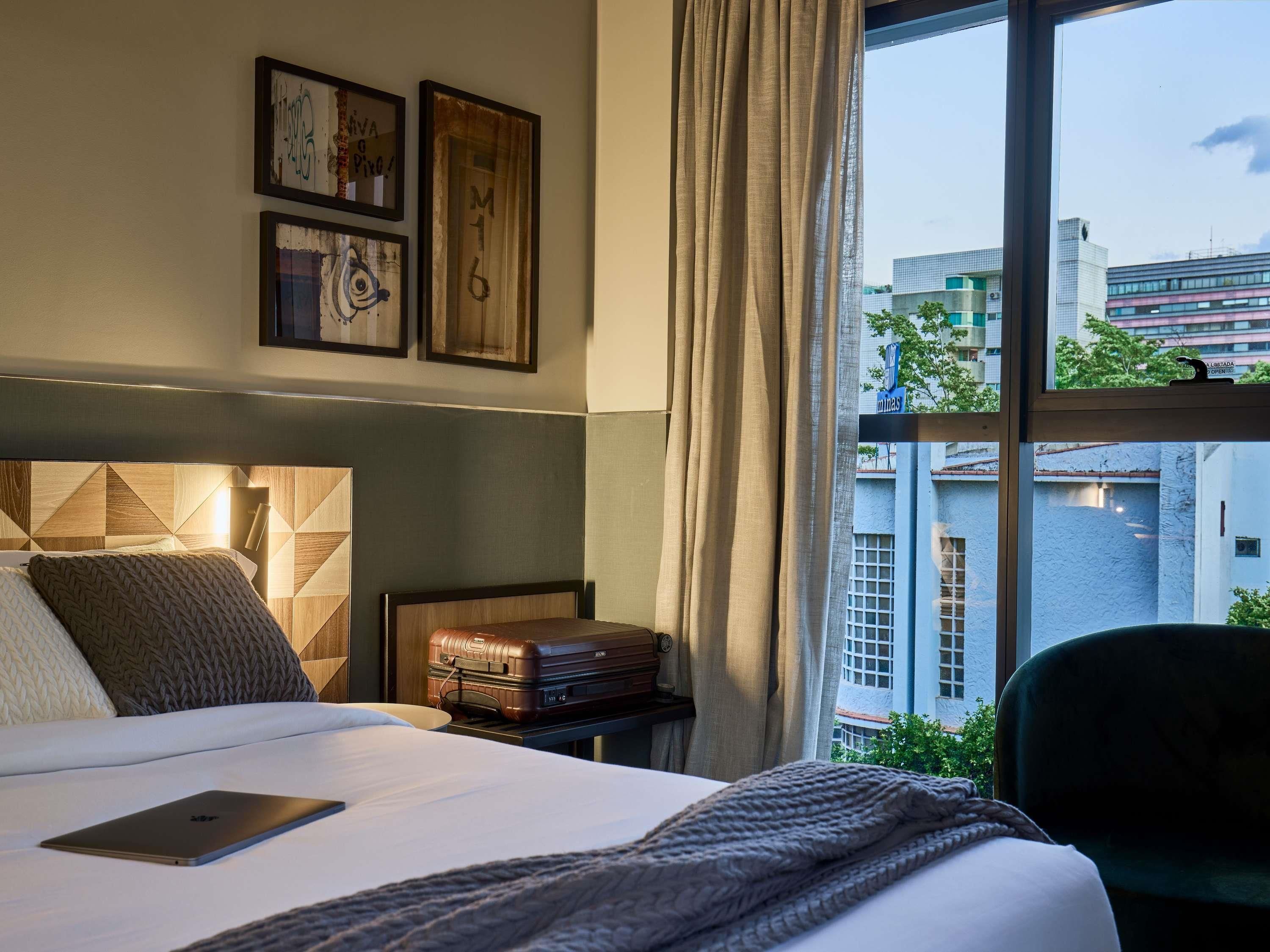 A hotel room with a bed, a suitcase, a laptop, and a view of buildings through the window.