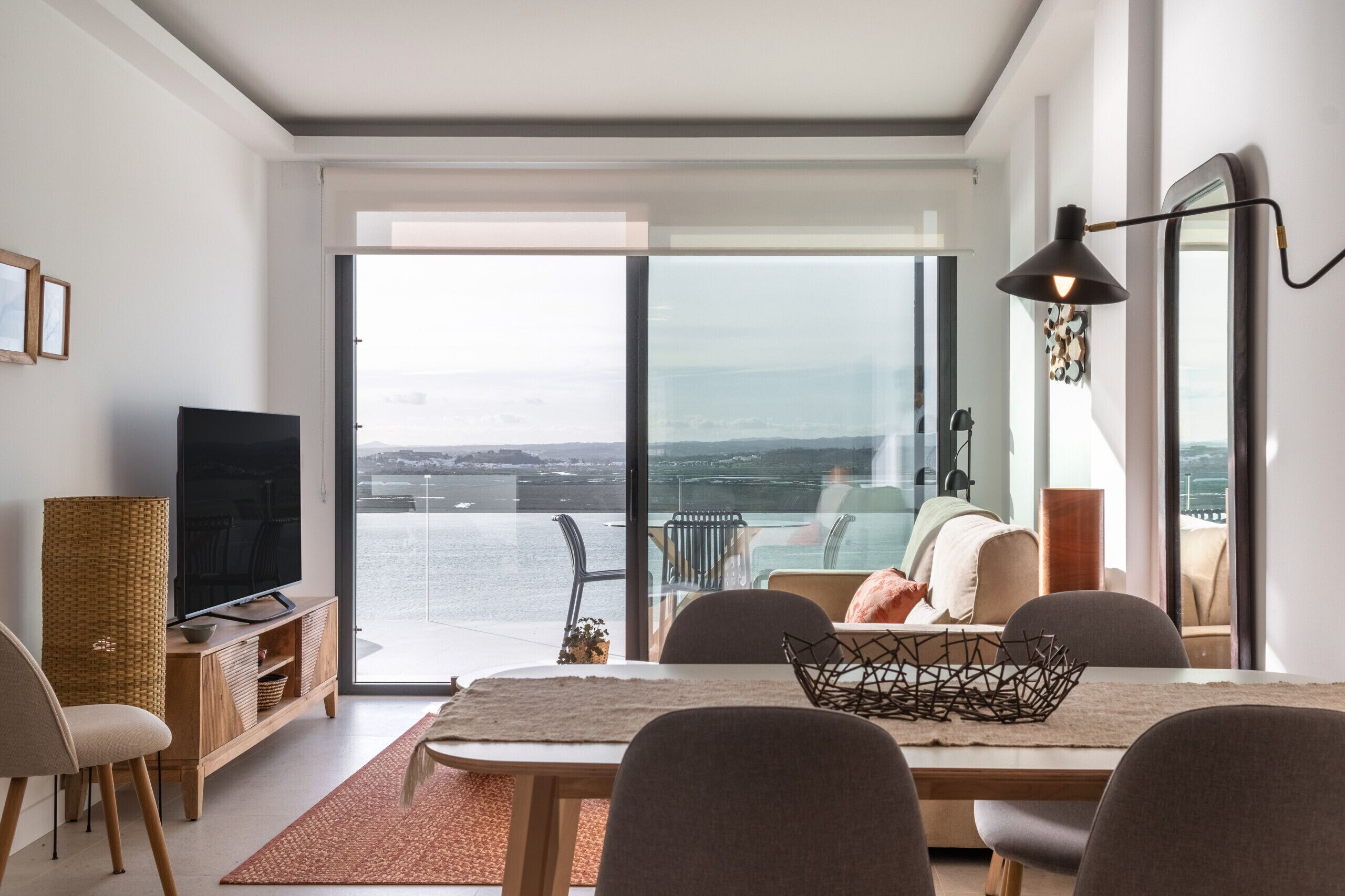 Un salon moderne doté d’une grande fenêtre donnant sur un plan d’eau, un coin repas avec une table et des chaises, et un téléviseur posé sur un meuble en bois.