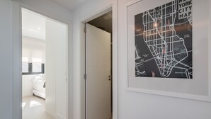 Un pasillo con una puerta blanca que da a una habitación con cama y ventana. En la pared hay un mapa enmarcado de la ciudad de Nueva York.