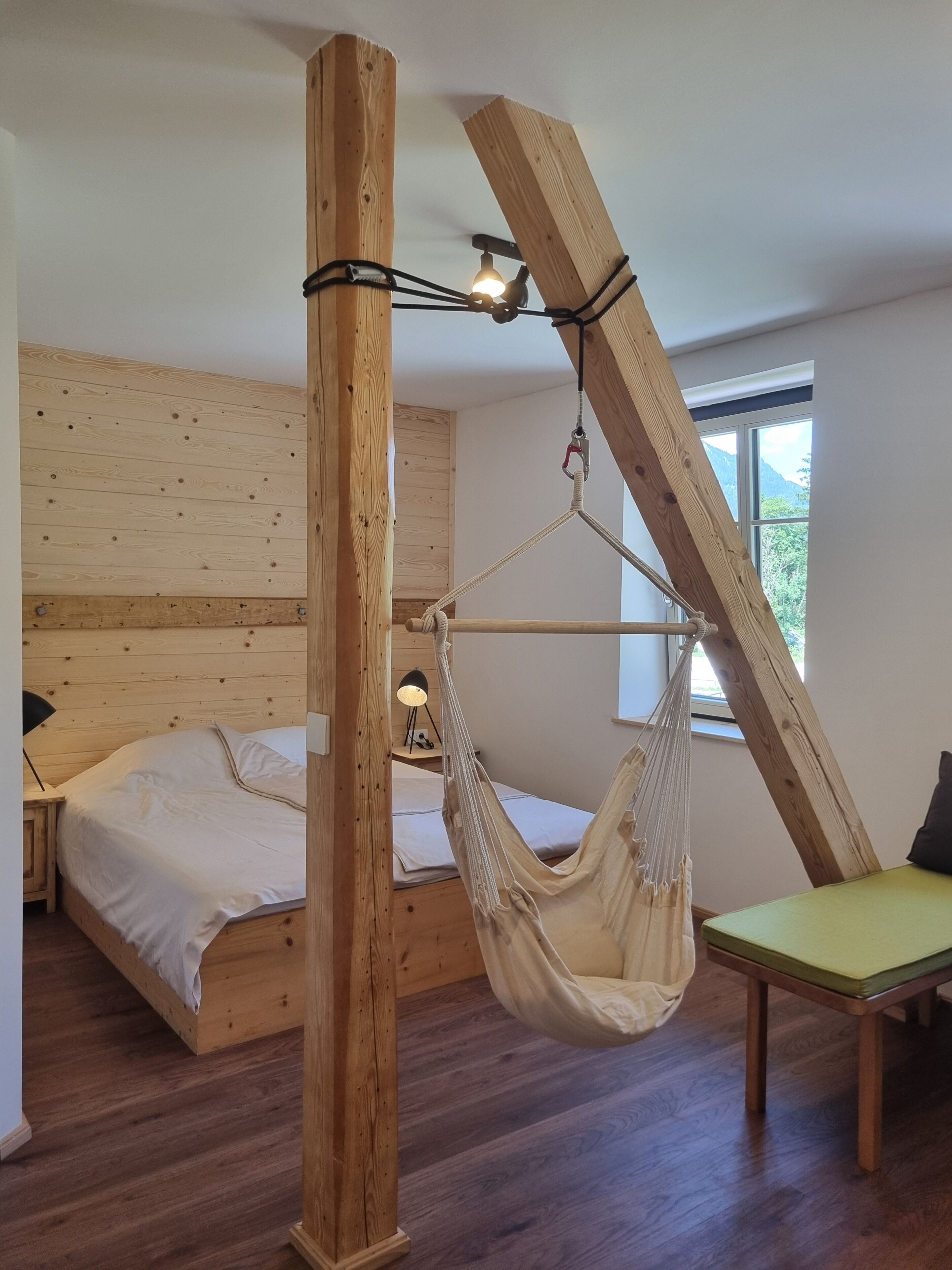 Una habitación con techo inclinado de madera, una cama con cubrecama blanco, una silla hamaca colgante y una mesita de noche con lámpara.