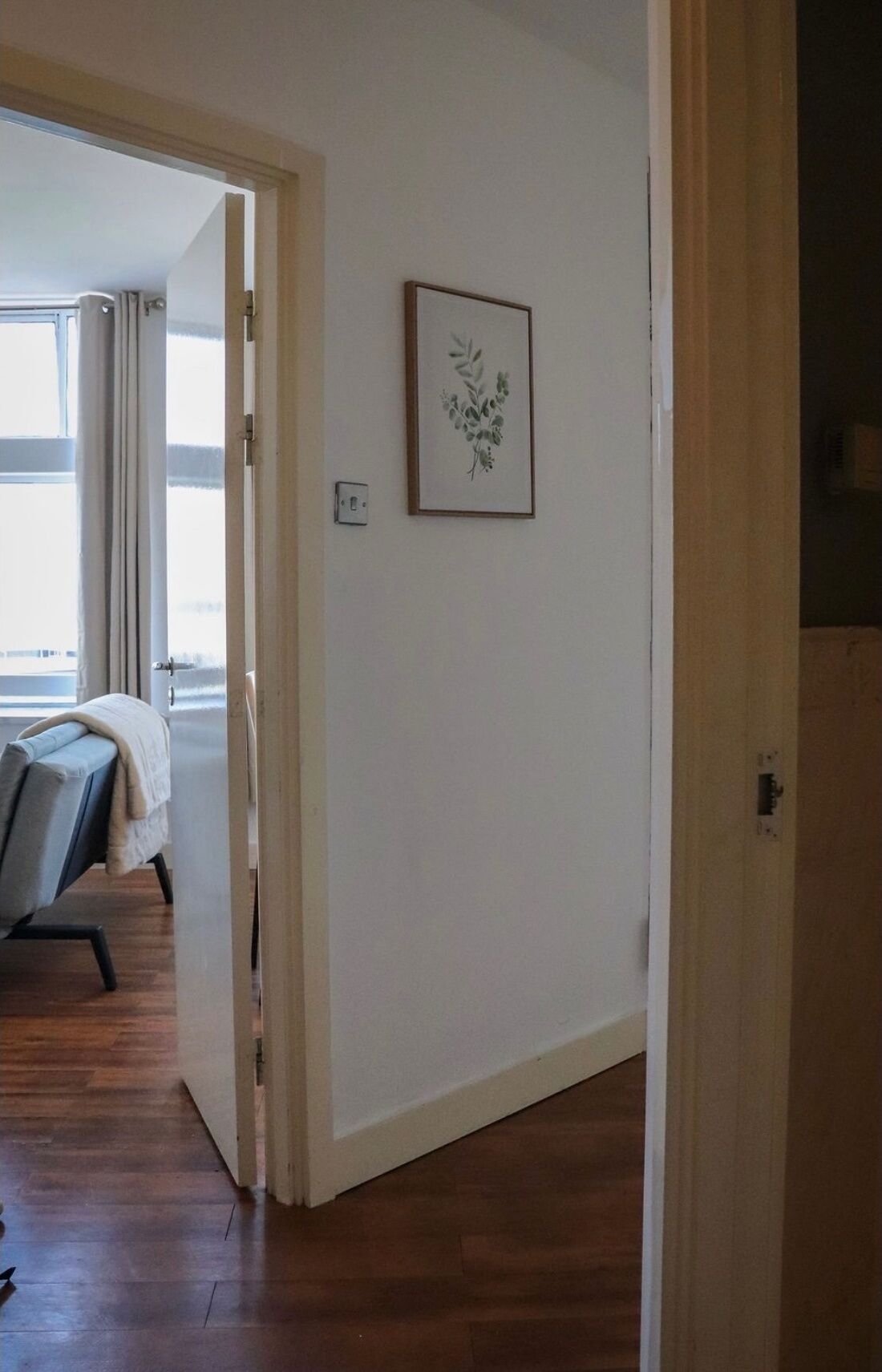 A hallway with a wooden floor leading to a room with a bed and a window.