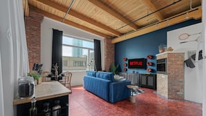 A modern kitchen with a blue sofa, a TV displaying 'NETFLIX', and a brick fireplace.