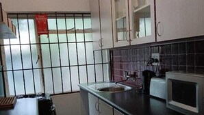 A kitchen with white cabinets, a microwave, a sink, and a window with bars. - Oaktree Guest House (Sandton)