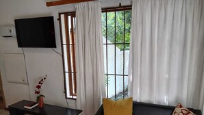 A living room with a black leather sofa, a yellow pillow, a black coffee table with a vase and book, a flat-screen TV on the wall, and a window with white curtains. - Oaktree Guest House (Sandton)