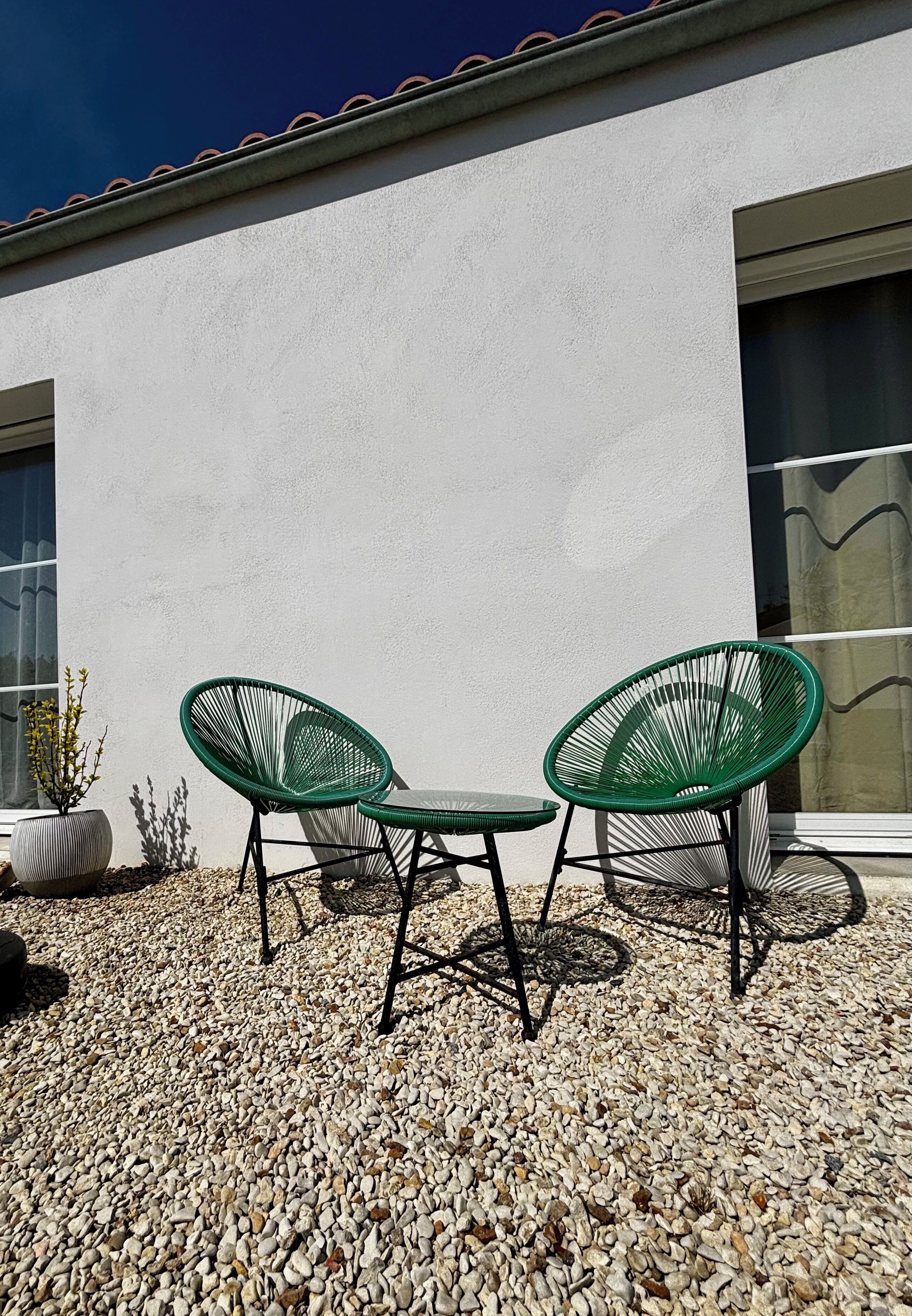 A patio with two green chairs and a table, set on pebbles, in front of a white building with a window and a door.