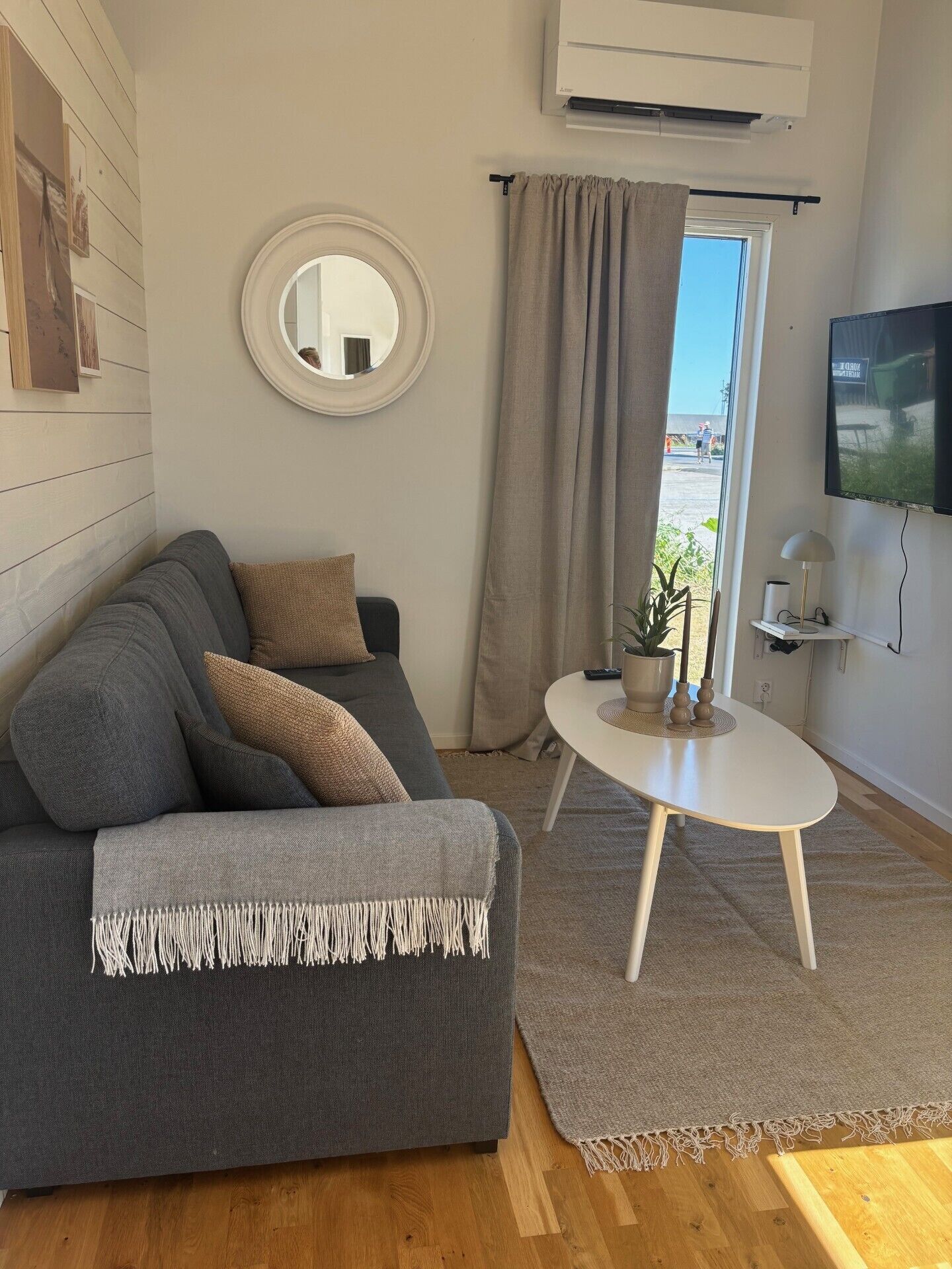 En moderne stue med en grå sofa, et rundt spejl, et fjernsyn og et lille bord med en plante.