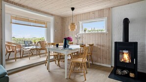 A cozy dining area with a white table and wooden chairs, a fireplace, and large windows offering a view of greenery. - Amazing home in Otterup with house sea view (Otterup)