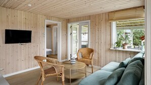 A cozy room with wooden walls and ceiling, a round dining table with chairs, a flat-screen TV mounted on the wall, and a window with a view of greenery. - Amazing home in Otterup with house sea view (Otterup)
