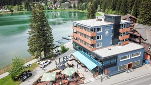 A multi-story building with a blue facade, a restaurant with outdoor seating, and a view of a lake. - Provisorium13 (Arosa)