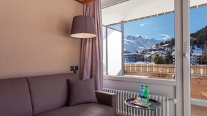 A room with a sofa, a table with bottles, and a view of mountains through a large window. - Provisorium13 (Arosa)