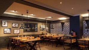 A dining area with multiple tables and chairs, pendant lighting, framed artwork on the walls, and a patterned tile backsplash. - Provisorium13 (Arosa)