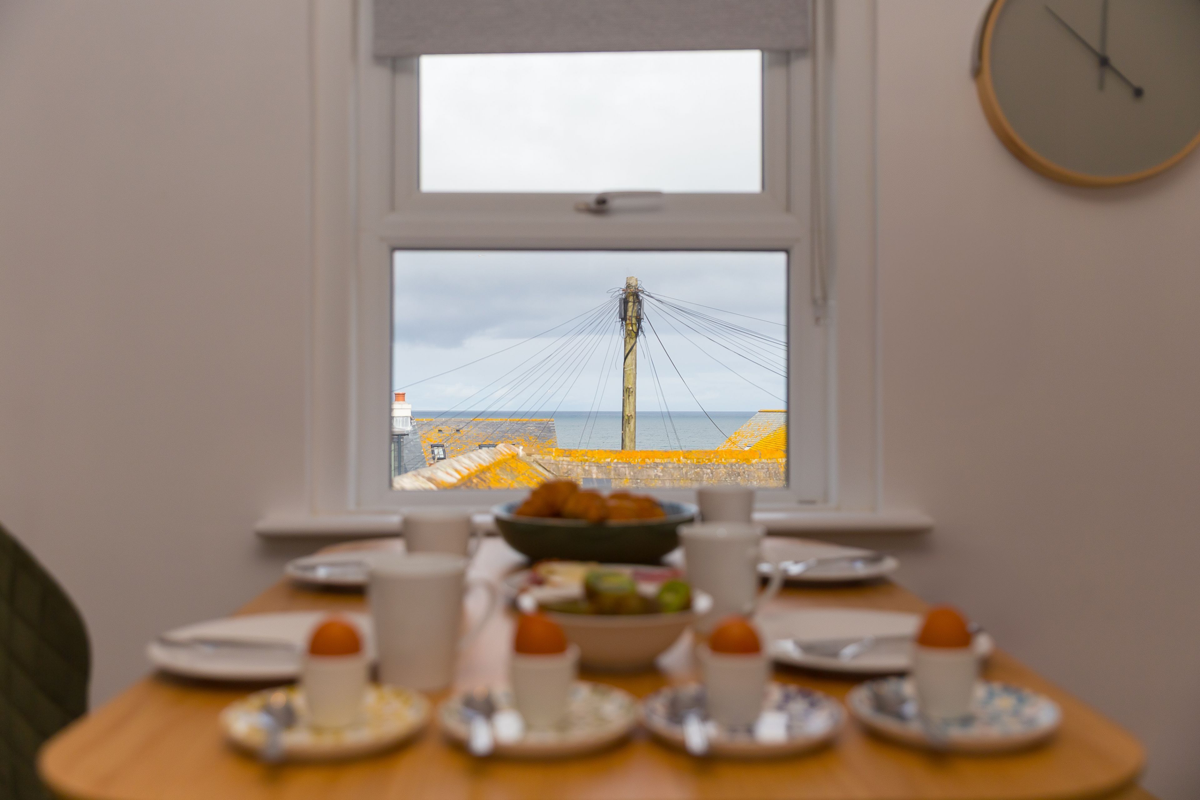 A breakfast table with plates, cups, and a bowl of fruit, set against a window overlooking a beach and power lines.
