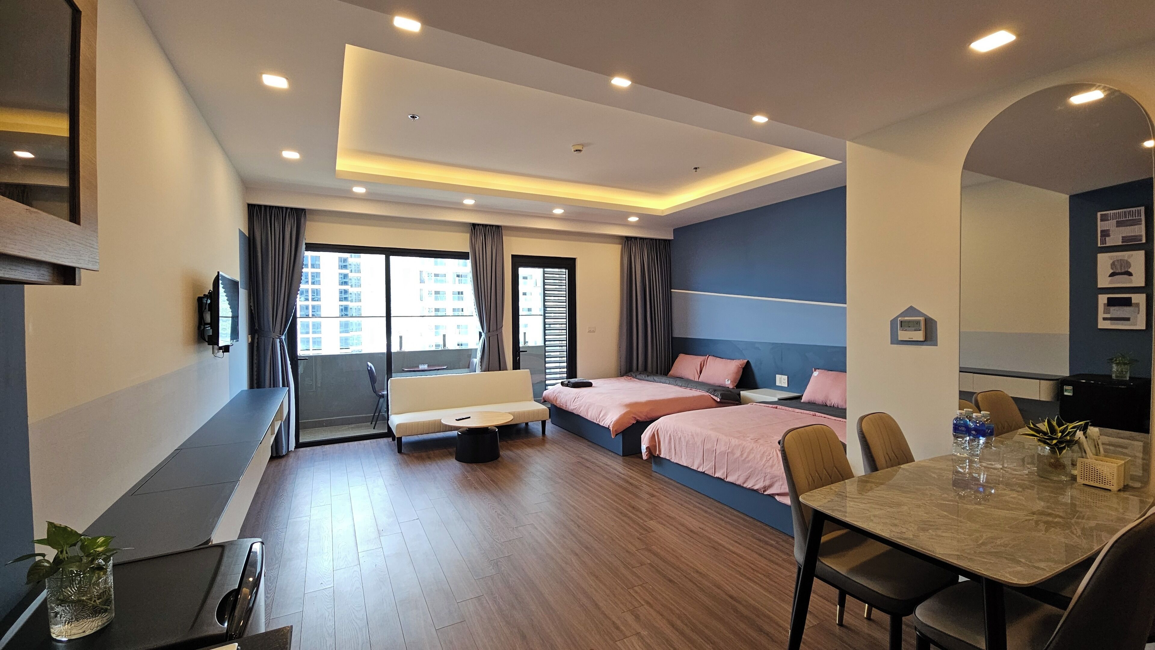 A modern hotel room with a sofa, two beds, a dining table, and a balcony with curtains.