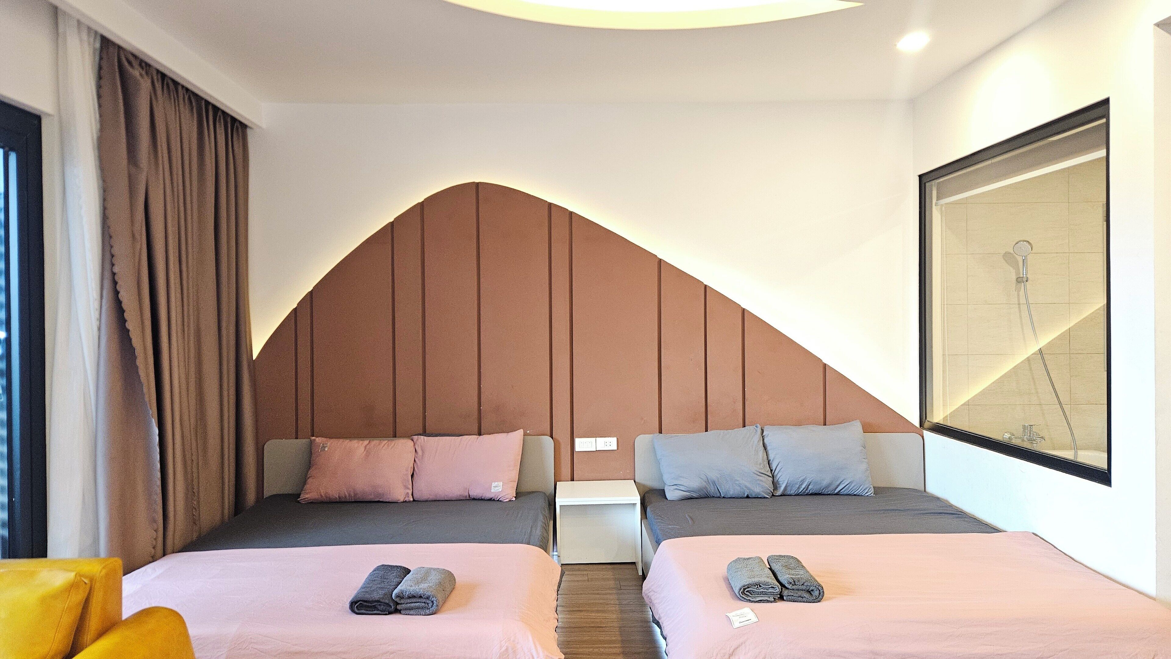 Hotellihuone, jossa on kaksi sänkyä, pieni yöpöytä, suuri peili ja suihkutila.