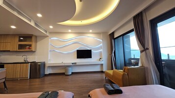 A modern hotel room with a curved ceiling, a flat-screen TV mounted on a wall, a sofa, a bed, a small kitchen area, and a large window offering a view.