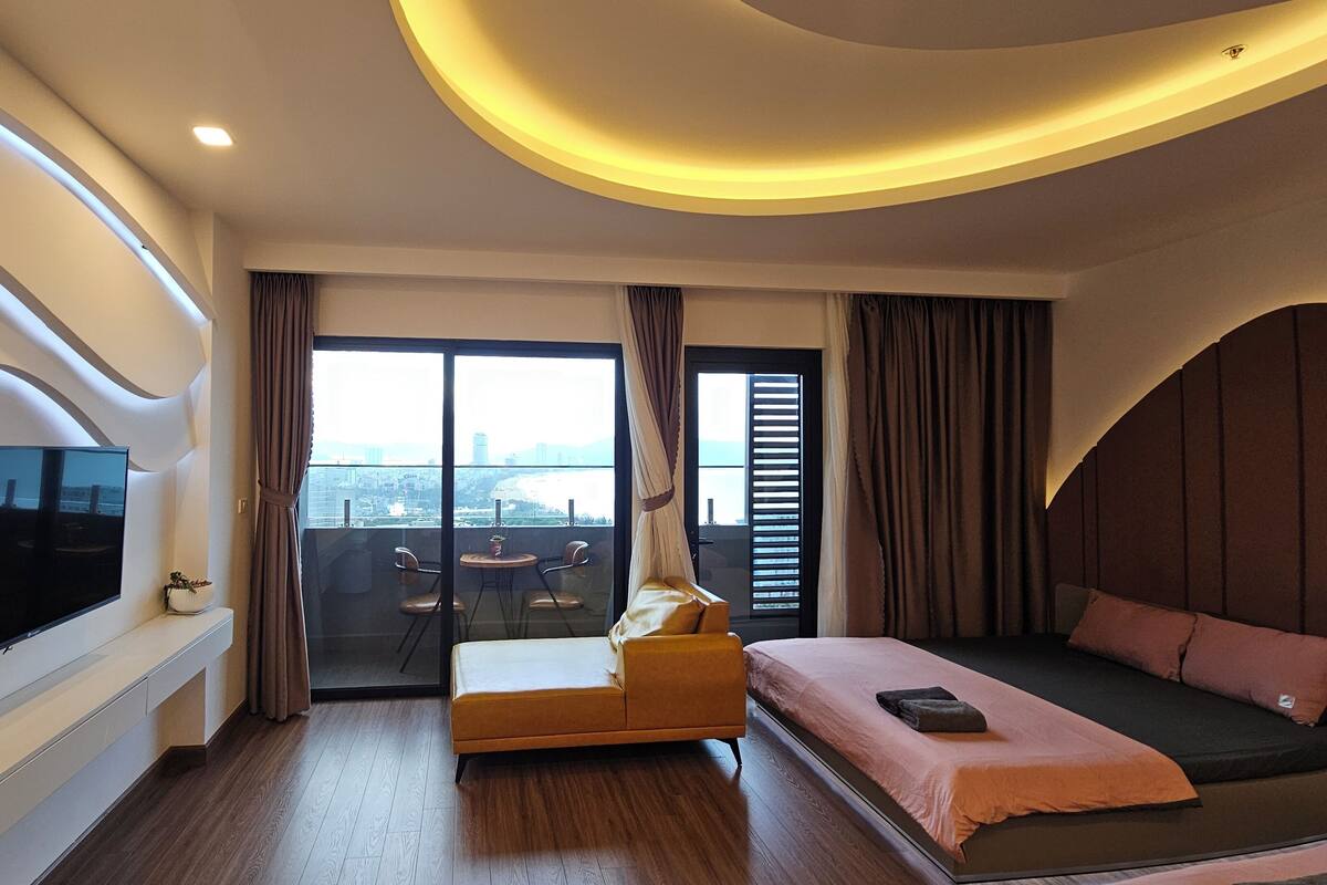 Habitación de hotel moderna con una cama grande, televisión, balcón y vistas a la ciudad, y un diseño de techo curvo.
