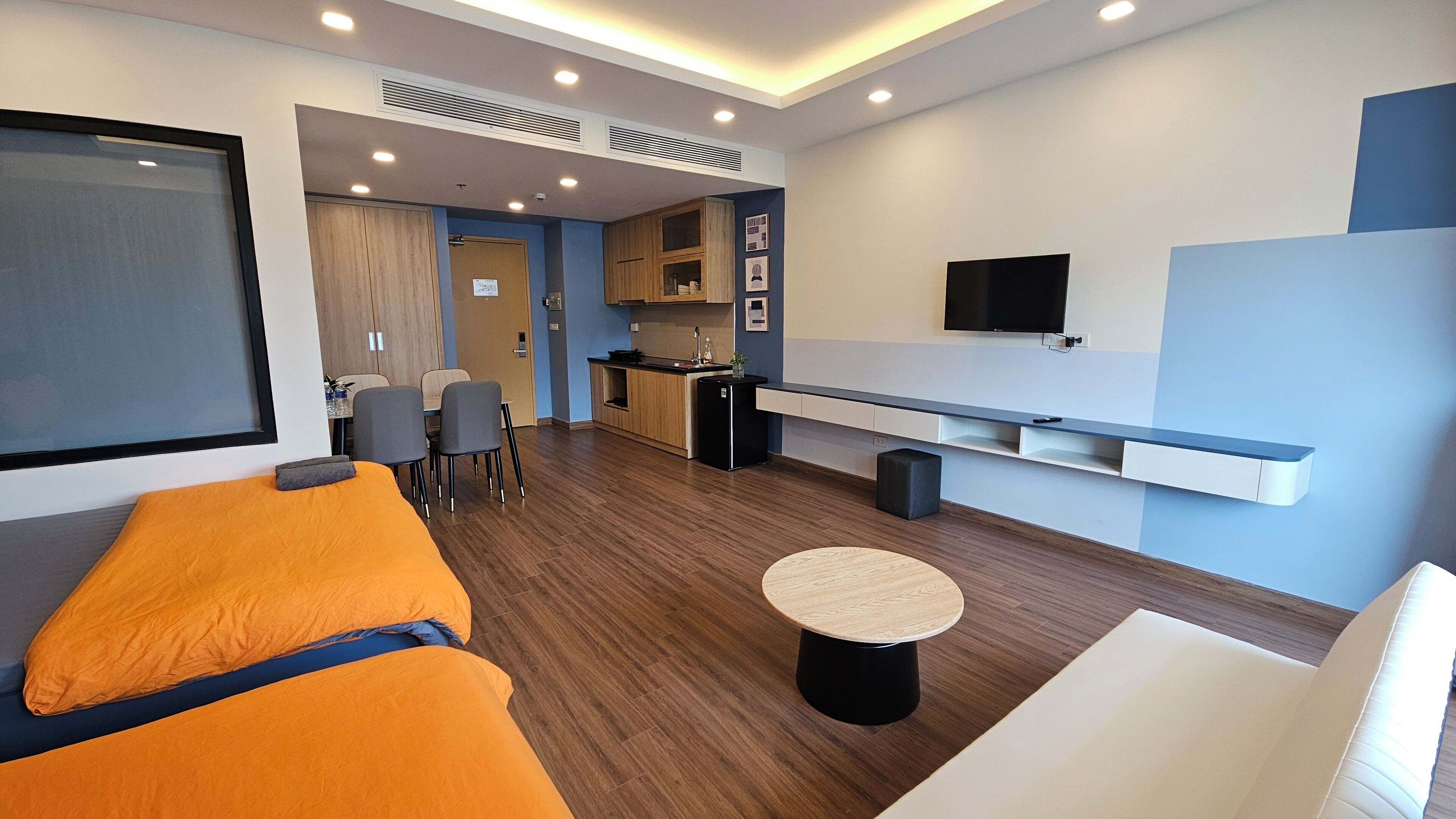A modern hotel room with a sofa, a small round table, a TV, a kitchenette, and a dining area.