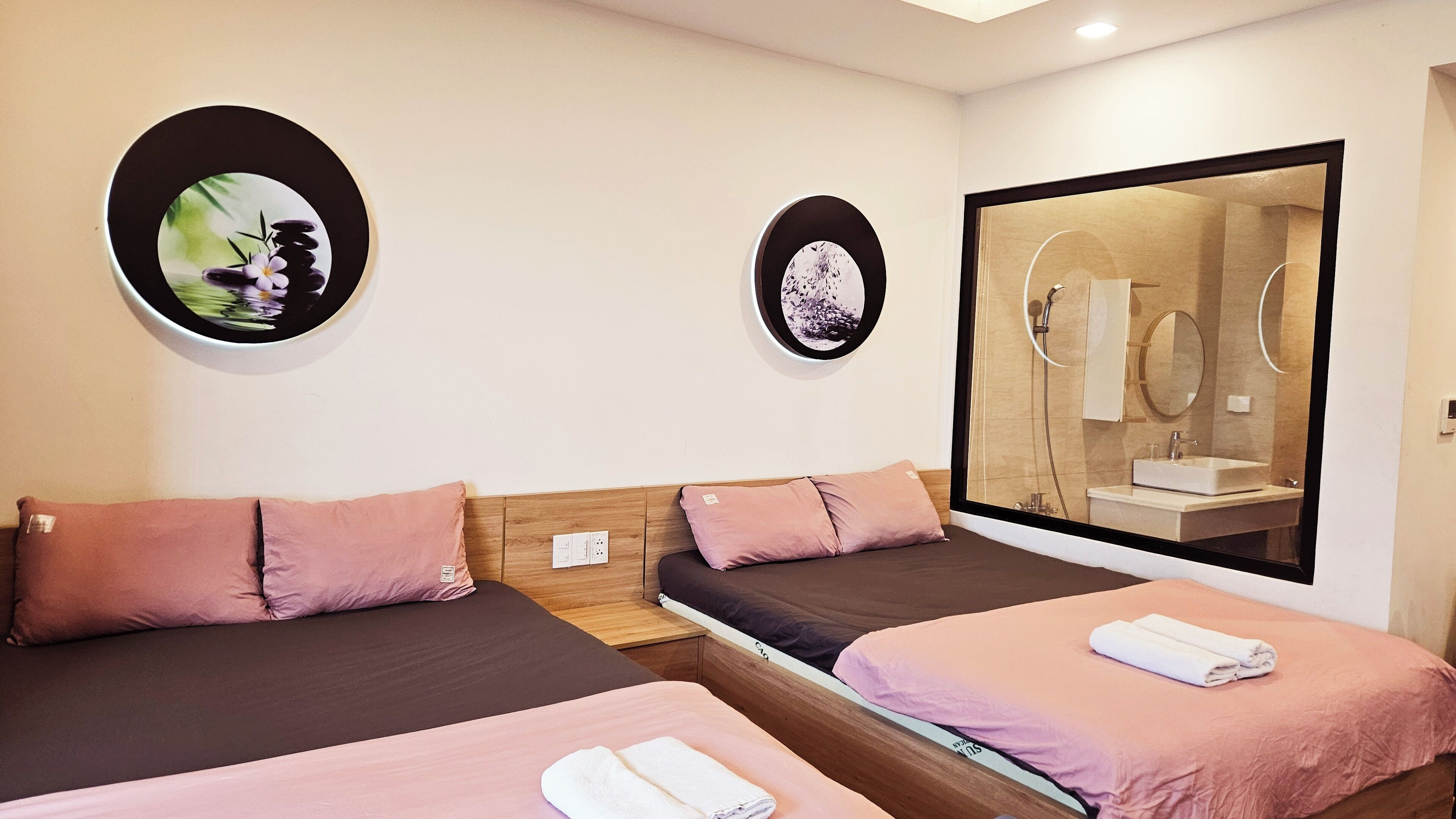 Une chambre d’hôtel avec trois lits, chacun agrémenté d’un oreiller rose et d’une serviette blanche. On y trouve deux miroirs ronds fixés au mur et un grand miroir qui reflète une salle de bain.