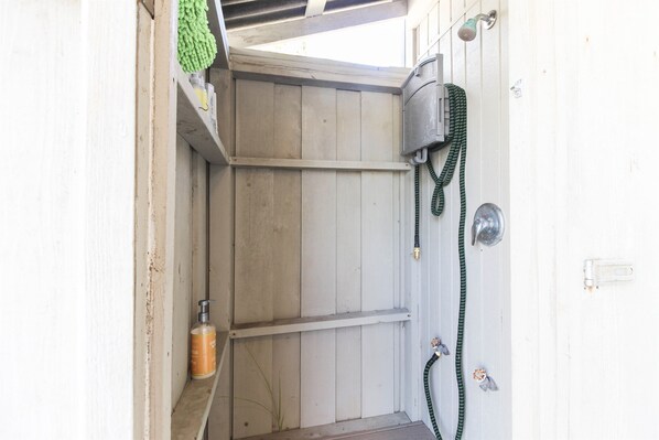 A small, enclosed outdoor shower area with a wooden bench, a green hose, and a metal showerhead.