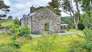 A stone house with a sloped roof, surrounded by lush greenery and a stone wall. - Penclogwynau (Dolgellau)