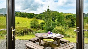 An outdoor wooden table with picnic setup, surrounded by a scenic view of green hills and a cloudy sky. - Penclogwynau (Dolgellau)
