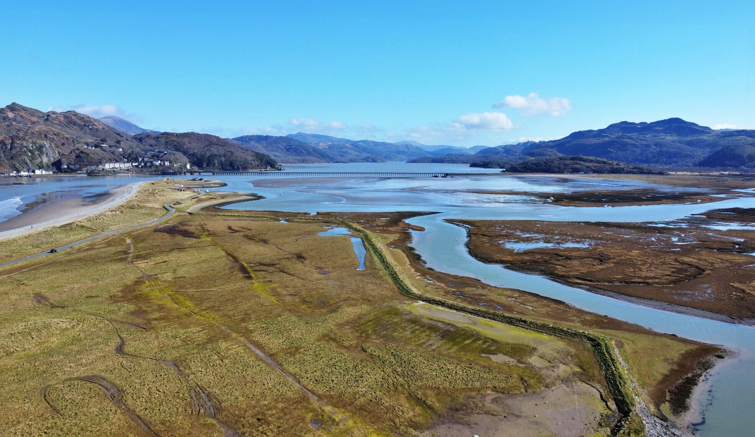 An aerial view of a coastal landscape with a bridge, a river, and mountains in the background.