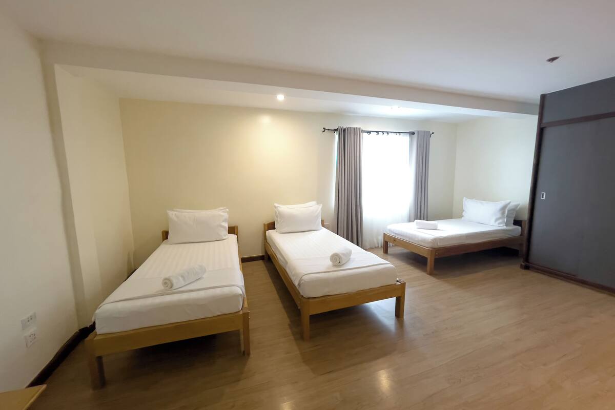 A hotel room with two single beds, a wooden floor, a grey wardrobe, and a window with curtains.