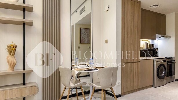 A modern kitchen with a built-in washing machine, a dining area with two chairs, and a large mirror reflecting the room. - Bloomfields Stylish Stu Burj View Arjan  (Dubai)
