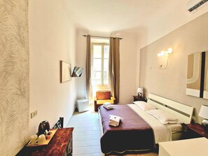 Una habitación de hotel con cama, mesita de noche, lámpara y vista al pasillo.
