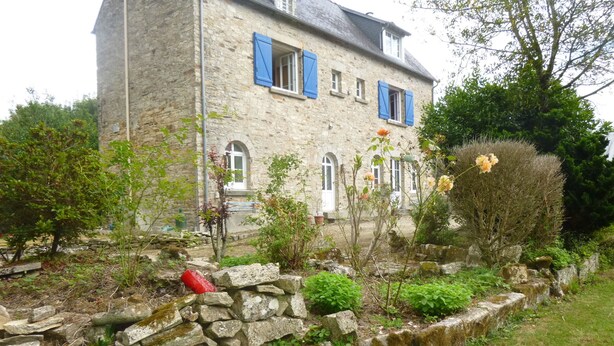 A stone house with blue shutters, a stone wall, and a garden with various plants and flowers.
