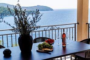 Una mesa con frutero, copa de vino y botella, con vistas a un paisaje de mar y colinas.