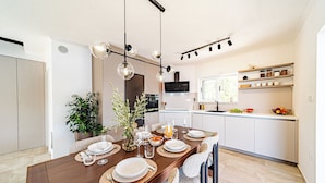 Un comedor moderno con una mesa de madera lista para una comida, rodeada de sillas. Se ve una cocina al fondo con armarios blancos y un horno empotrado.