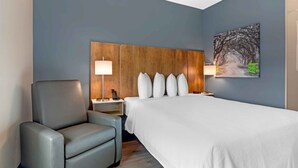 Hotellihuone, jossa on suuri sänky, puinen pääty, kaksi yöpöytävalaisinta, harmaa nojatuoli ja taulu seinällä.