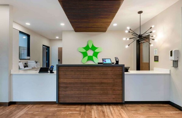 A reception area with a wooden reception desk, a green logo, a modern chandelier, and a clean, minimalist design.