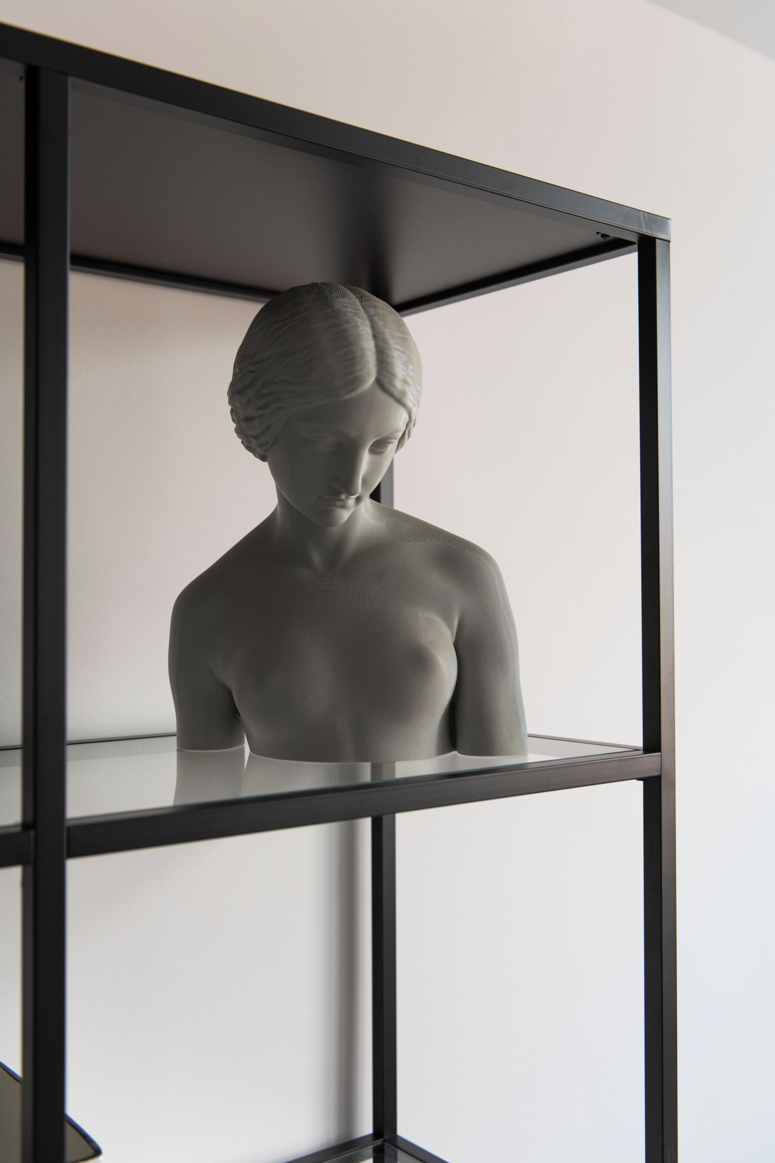 A sculpture of a human figure on a black metal shelf against a plain wall.