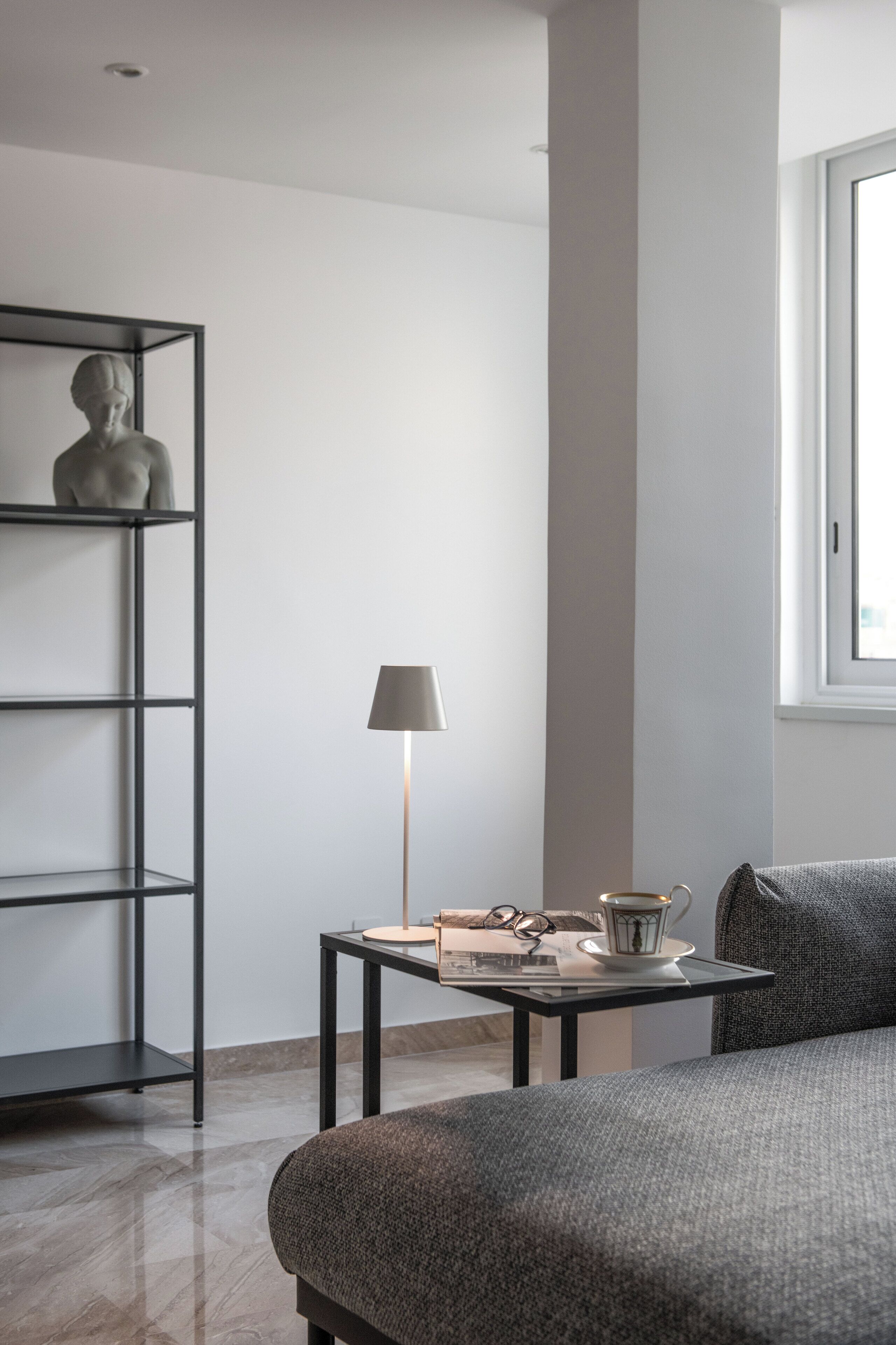 A modern bedroom with a bed, a bedside table with a lamp, a small shelf with a statue, and a window providing natural light.