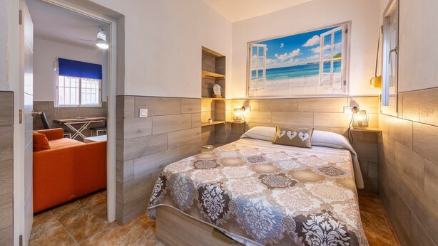 Un dormitorio con cama, mesita de noche, lámpara de pared, un cuadro de una playa y vistas a un balcón.
