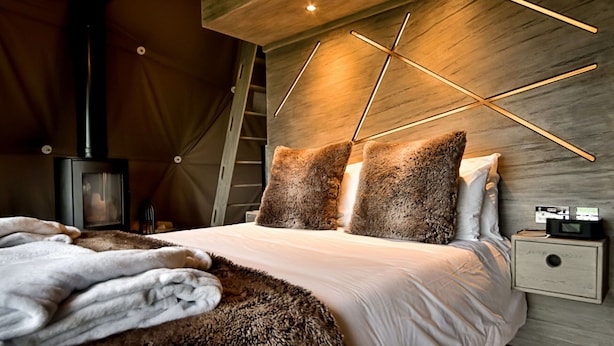 A modern bedroom with a wooden ceiling, a bed with fluffy pillows, a fur throw, a nightstand with a radio, and a wood-burning stove.