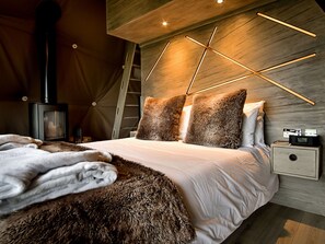 Ein modernes Schlafzimmer mit Holzboden, einem Bett mit flauschigen Kissen, einer Felldecke, einem Nachttisch mit Radio und einem Kaminofen.
