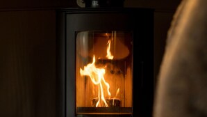 A wood-burning stove with visible flames inside a glass enclosure, set against a wooden wall.
